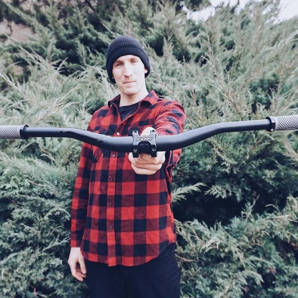 A person in a red and black plaid shirt and black beanie stands outdoors in front of green bushes, holding a Funn bicycle handlebar toward the camera.