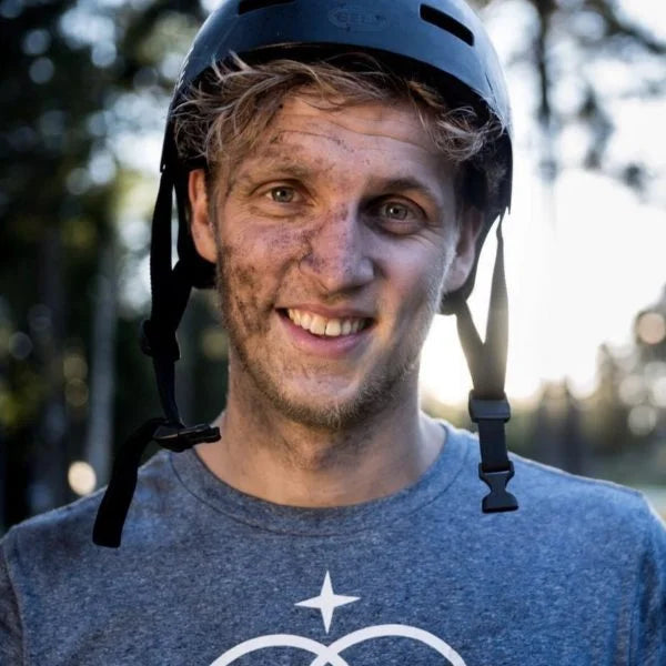 A smiling young man wearing a helmet and a grey t-shirt, with dirt on his face, stands outdoors with trees and sunlight in the background.