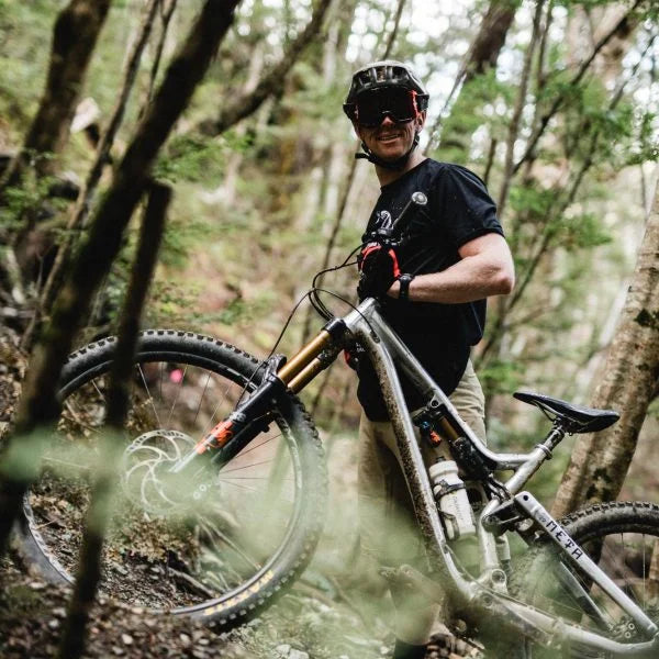 A mountain biker wearing a helmet, Funn sunglasses, gloves, and a black shirt stands smiling next to his mountain bike on a wooded trail surrounded by trees.