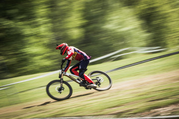 A cyclist in red and purple gear rides a mountain bike at high speed on a dirt trail through a green, blurry forest background, conveying a sense of motion.