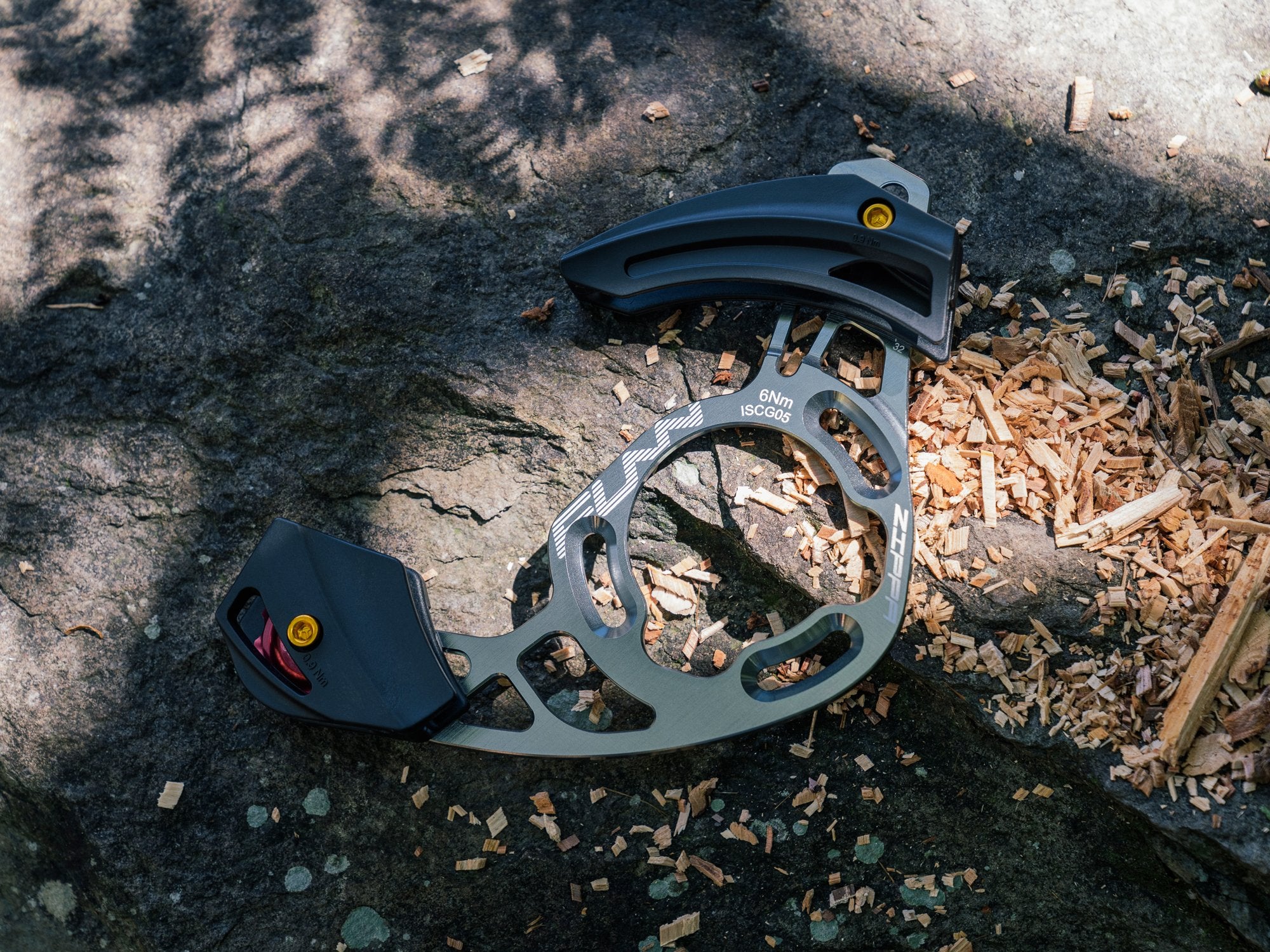 A close-up of a gray funn zippa am mountain bike chain guide resting on a rock surface, surrounded by wood chips and illuminated by dappled sunlight.