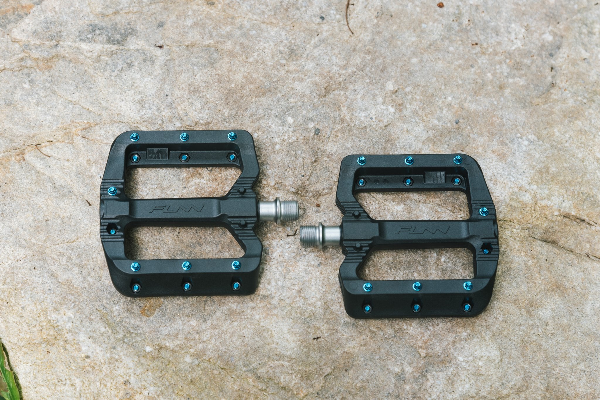 A pair of black bicycle pedals with blue screws, placed side by side on a rough stone surface.
