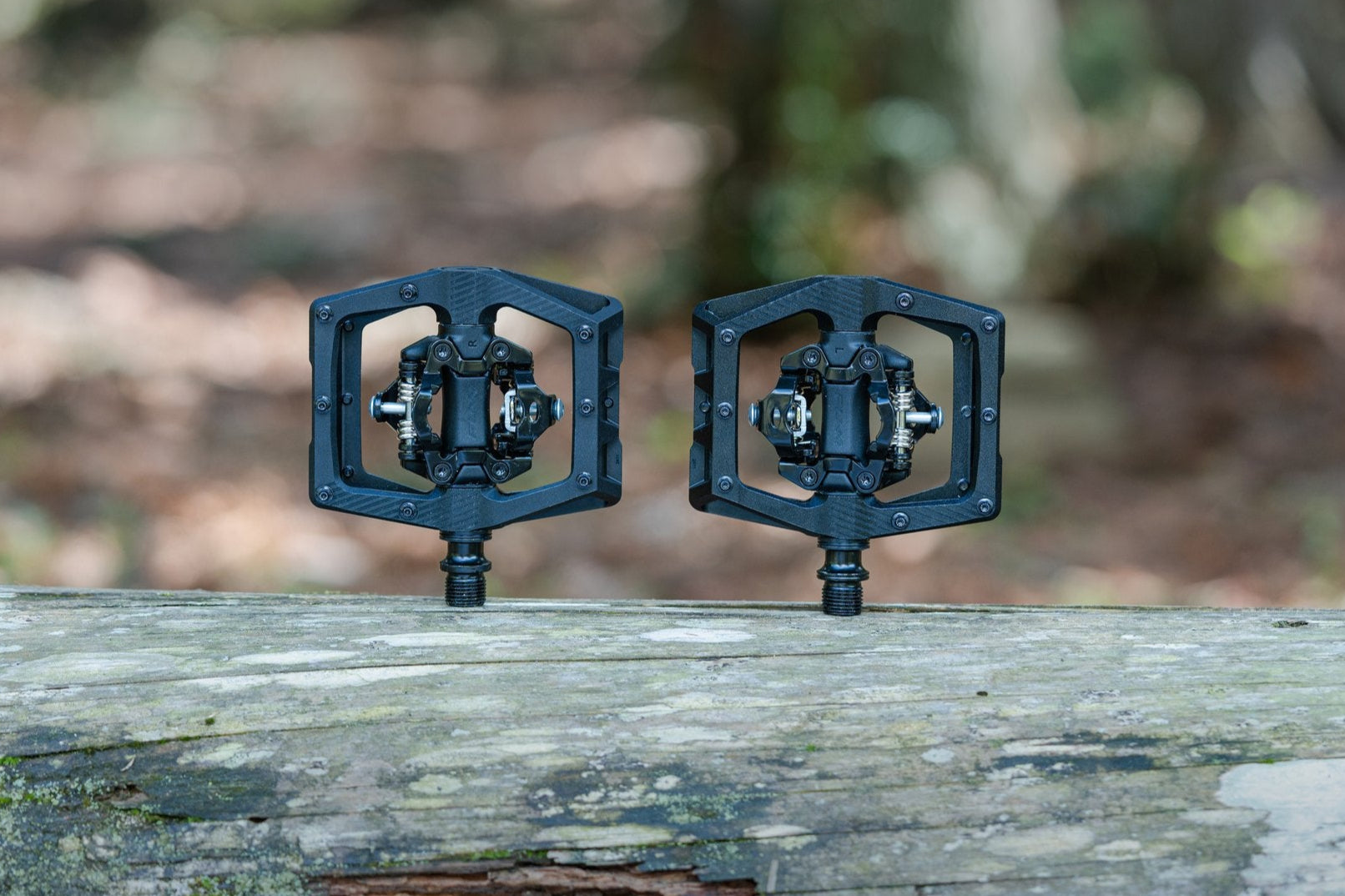 Two black metal bicycle pedals with open hexagonal frames are resting side by side on a weathered log, with a blurred forest background.
