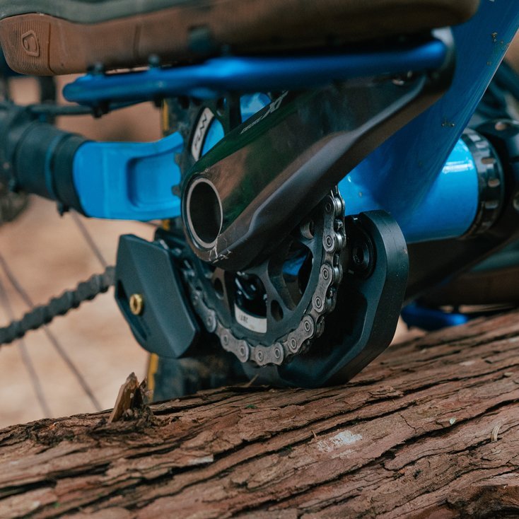 Close-up of a blue mountain bike crank and chainring resting on a fallen tree, highlighting drivetrain components and the Funn Zippa LG Chain Guide with its CNC-machined alloy backplate.