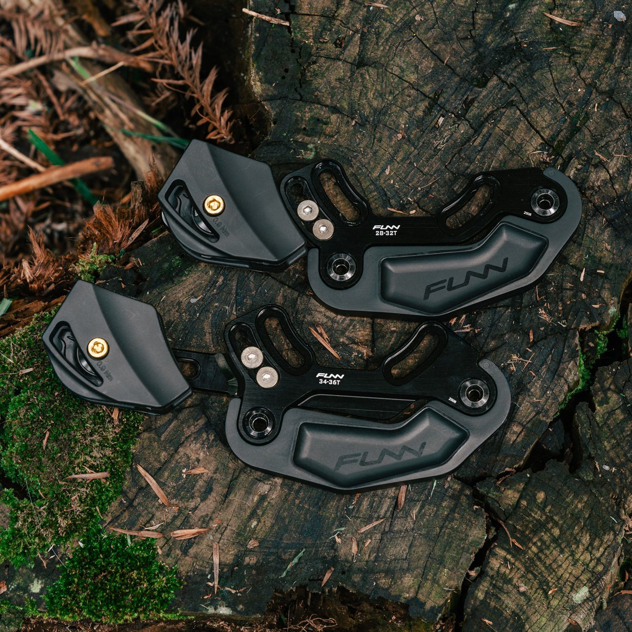 Two black Funn Zippa LG Chain Guides, CNC-machined from alloy, rest on a mossy tree stump surrounded by greenery and leaves in a forest—perfect for high-pivot bikes.
