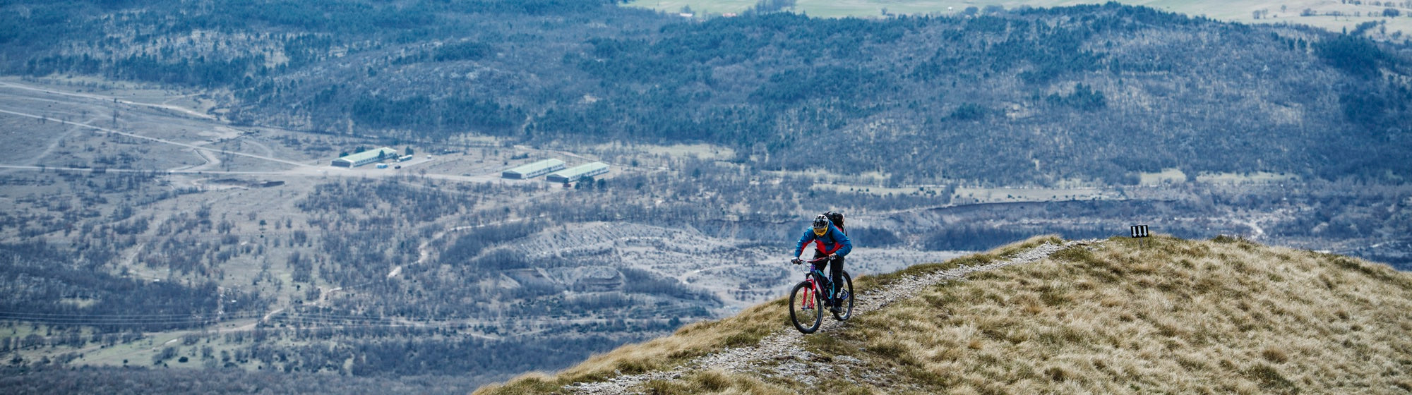 A person wearing a helmet and outdoor gear rides a Funn-equipped mountain bike along a grassy ridge, with a vast, hilly landscape stretching out in the background.