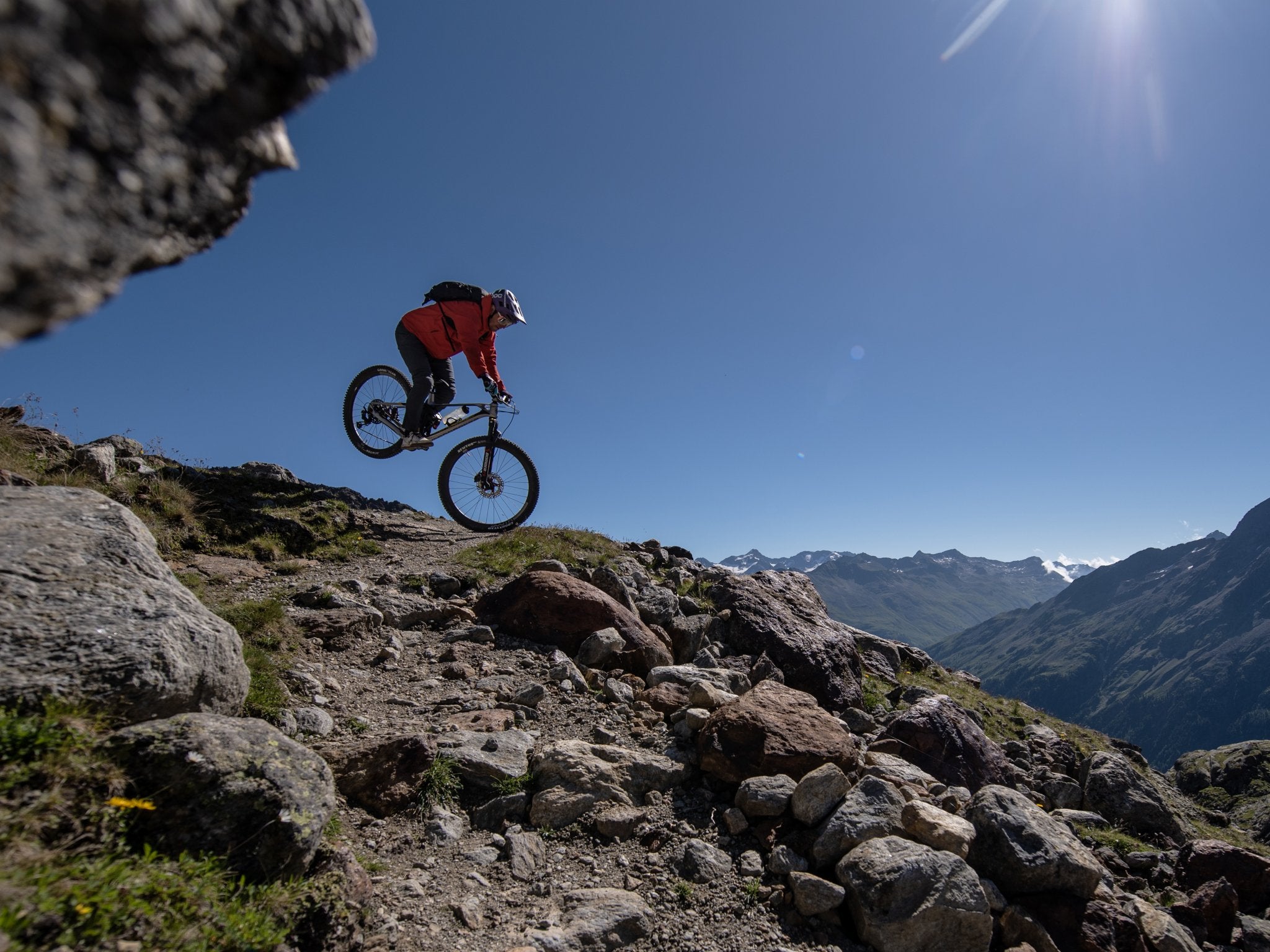 A mountain biker in a red jacket is mid-air, jumping over a rocky trail on a high-altitude mountain, with a stunning backdrop of distant peaks under a clear blue sky.