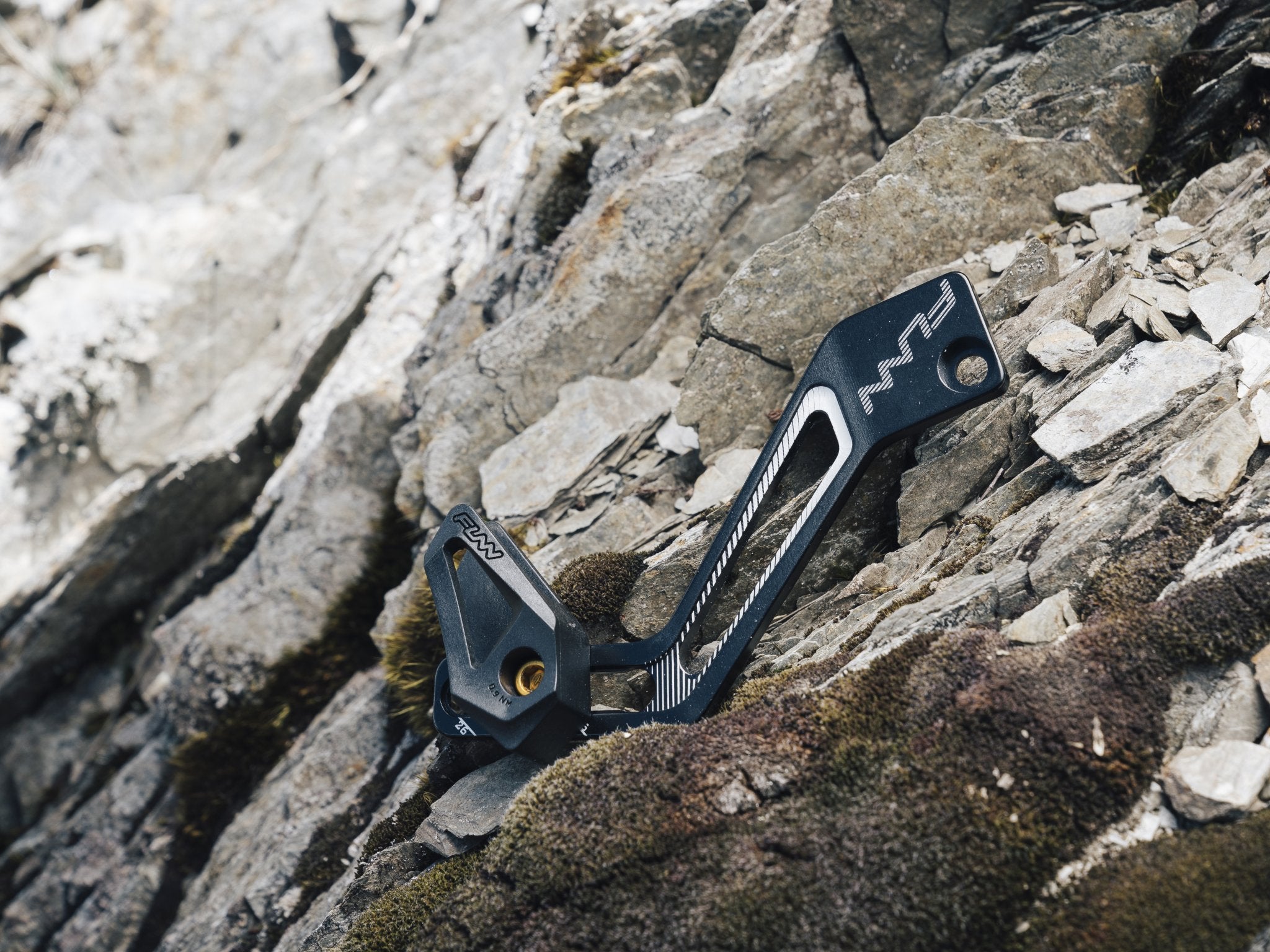 A close-up of a Funn zippa lite d-type chain guide resting on rugged, moss-covered rocks in an outdoor setting.