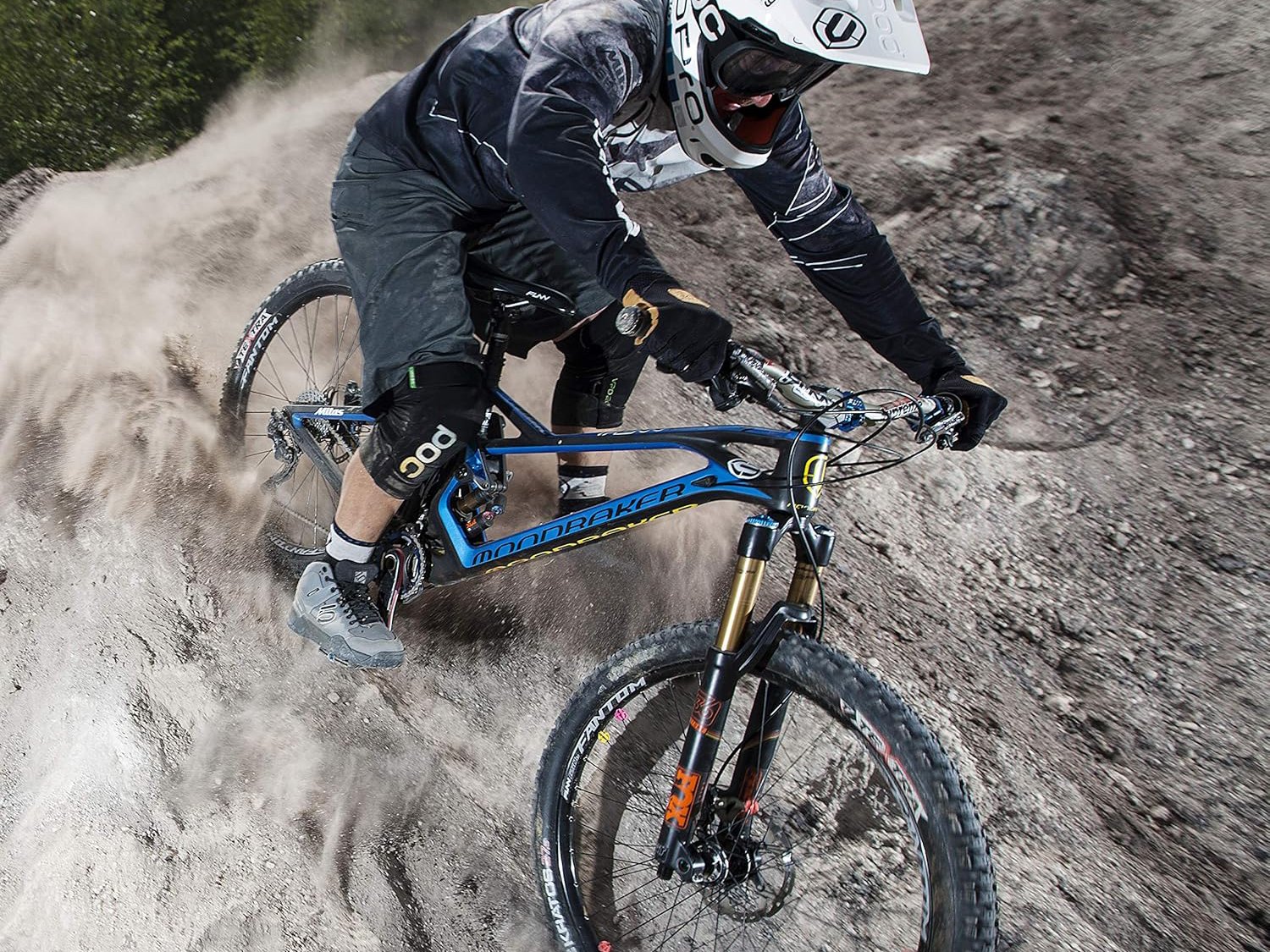 A funn mountain biker, Gaspi, in full gear descends a steep, dusty trail, kicking up dirt on a blue full-suspension bike.