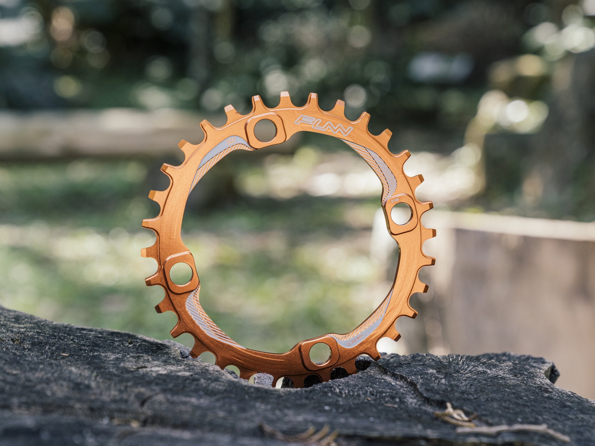 This image features an orange Funn solo 104 bcd chainring standing vertically on a rugged surface, set against a blurred outdoor background. 