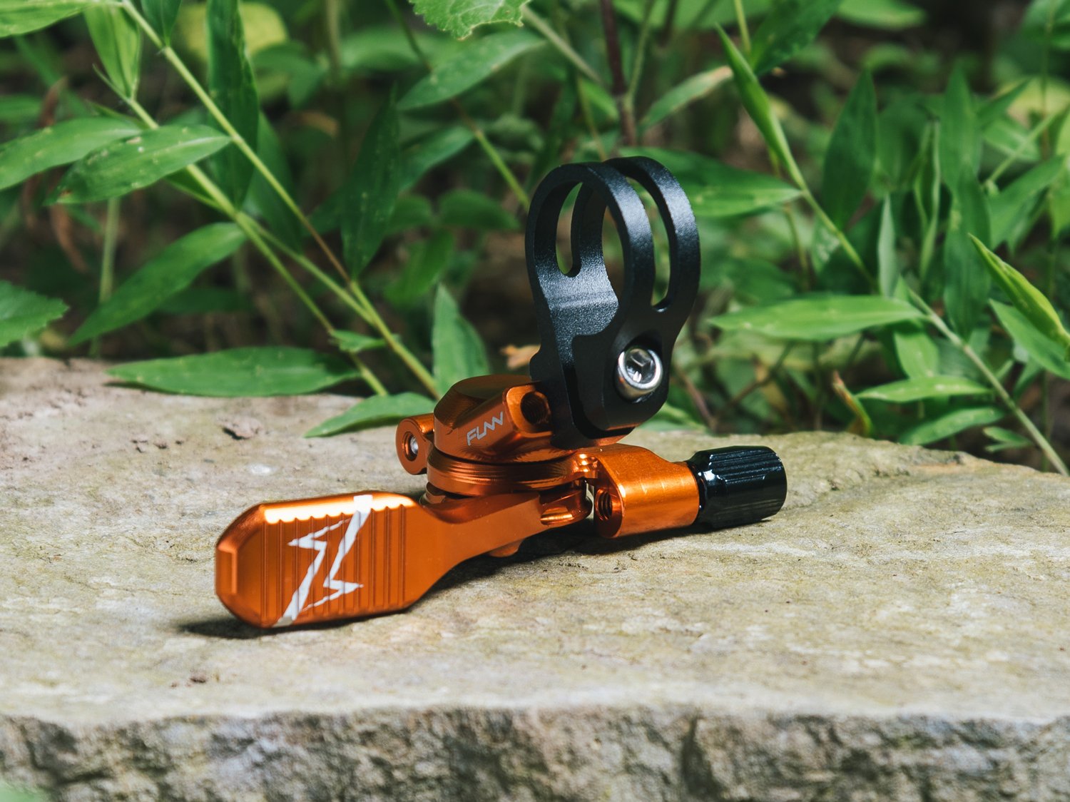 This image shows a vibrant orange funn updown anodized dropper post remote lever, placed on a natural stone surface with greenery in the background.