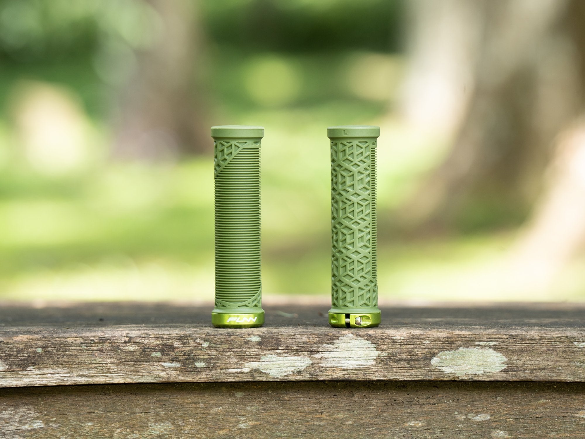 A pair of olive green funn hilt jr mountain bike grips, each with a different texture pattern, displayed side by side on a wooden surface with a blurred natural background.