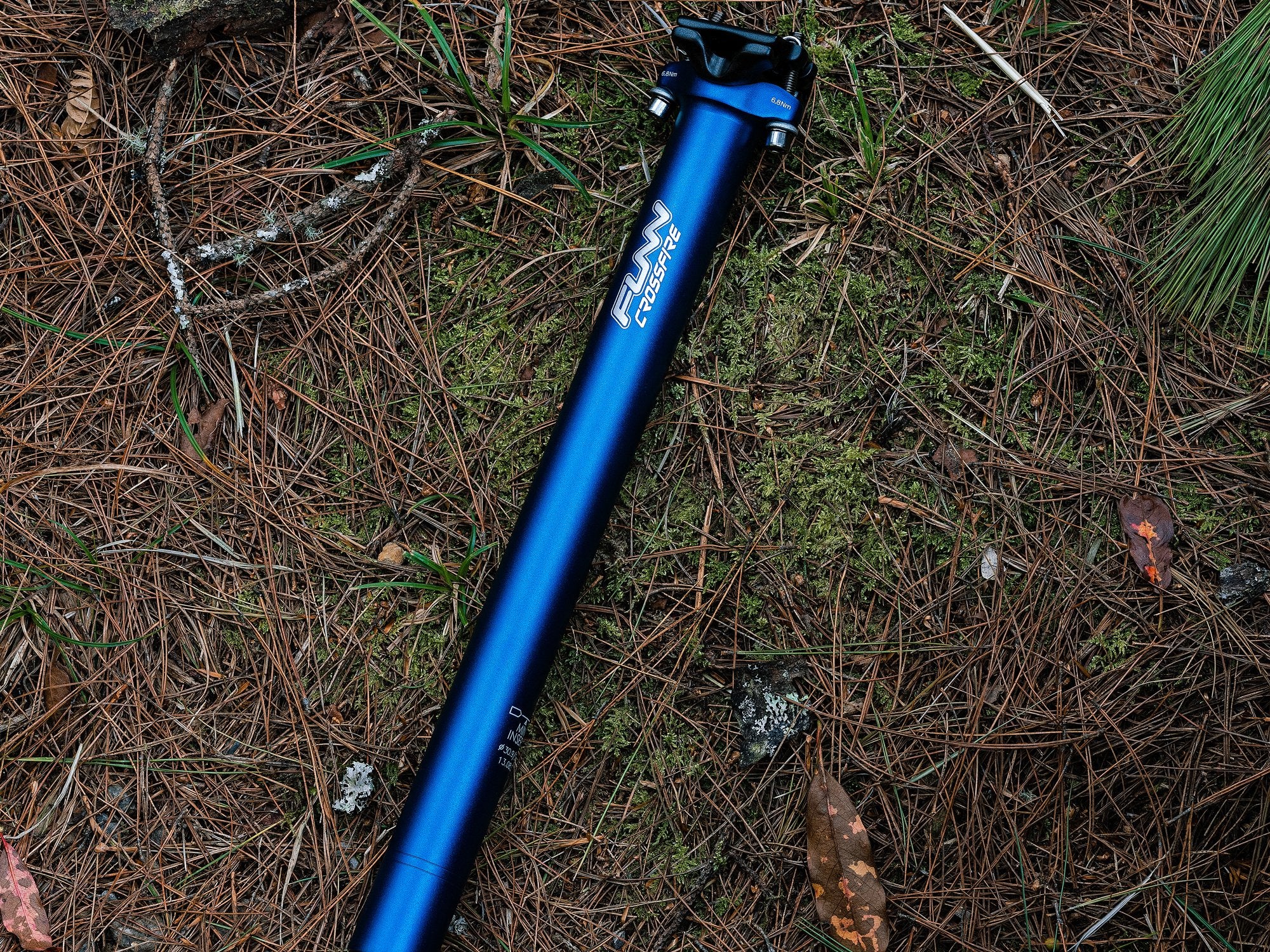 The image displays a blue Funn crossfire seat post arranged in a row on a forest floor covered with pine needles.