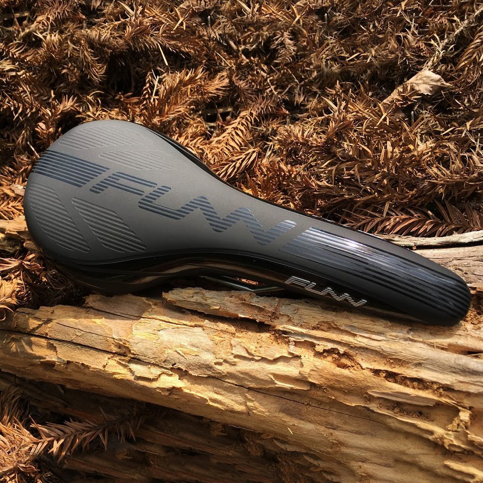 Black FUNN Adlib Saddle bike seat with sleek ergonomic shape and textured surface, resting on outdoor trail logs.