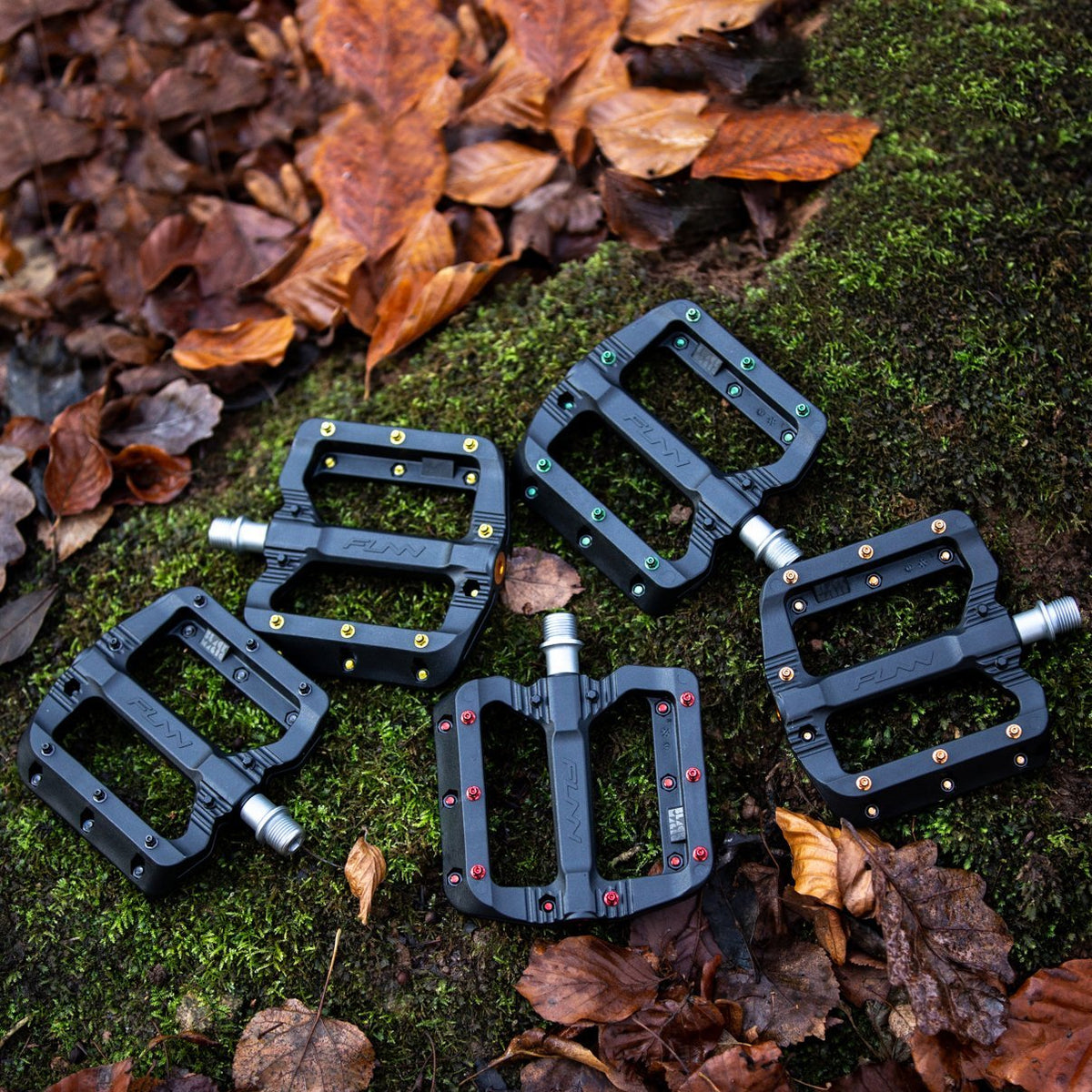 FUNN Black Magic Flat Pedals in durable composite, wide black platform with colored pins for MTB grip, displayed on forest moss