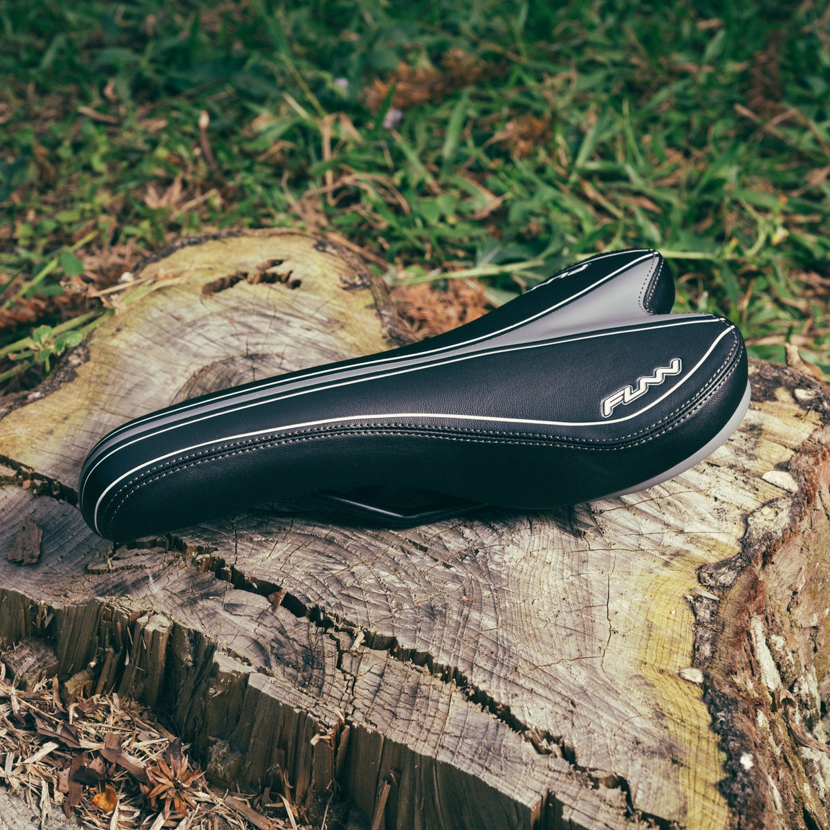 FUNN Launch II Saddle, black synthetic ergonomic bike seat with white stitching and logo, resting outdoors on a tree stump
