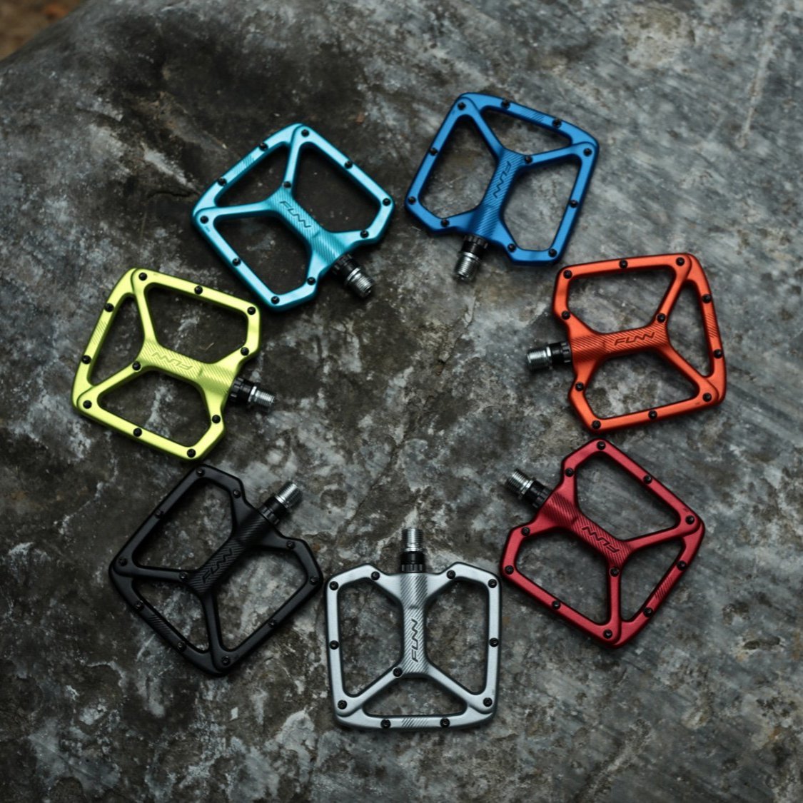 Colorful Funn Python flat pedals in anodized alloy, wide platform with traction pins, arranged in a circle on rocky ground