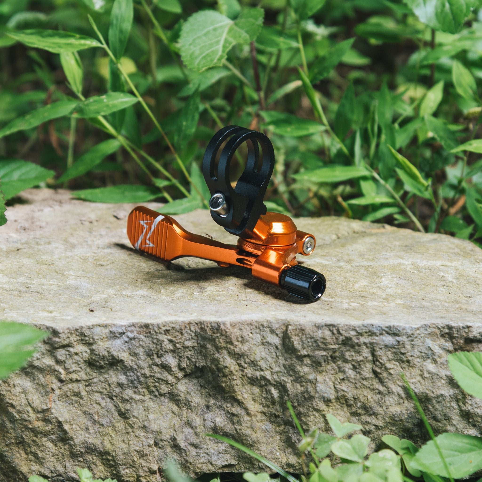 FUNN UpDown Dropper Lever, orange alloy dropper seatpost remote with black clamp, shown outdoors on a rock among greenery