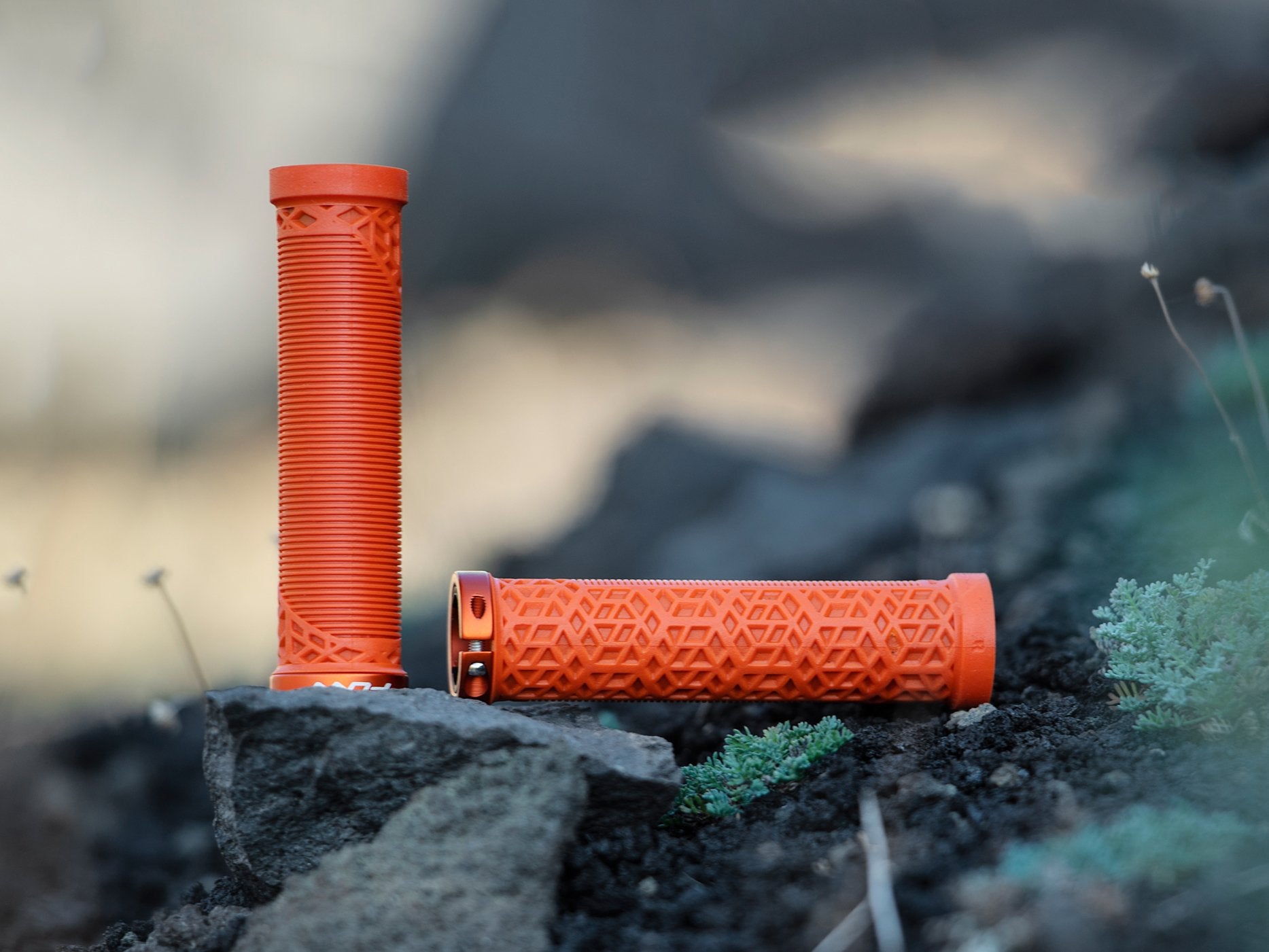 A pair of orange funn hilt mountain bike grips, one standing upright and the other lying down, displayed on rocks and surrounded by greenery in a natural outdoor setting.