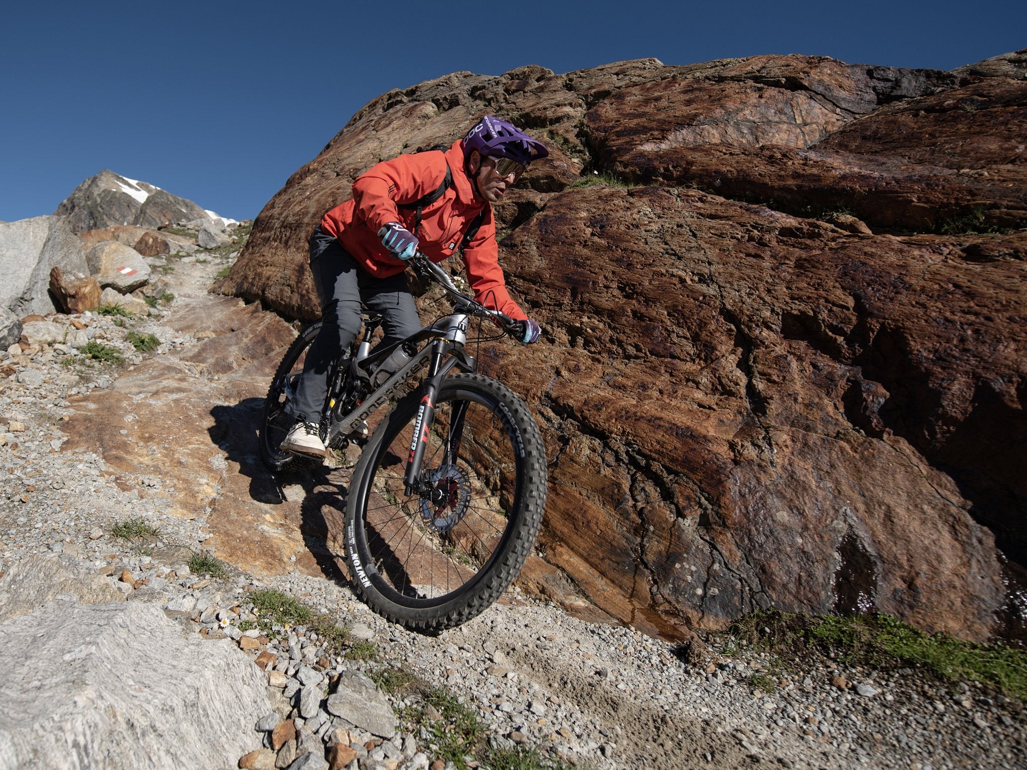 A mountain biker in a red jacket and purple helmet rides along a rocky trail, descending past large boulders on a technical section with a clear blue sky and distant mountains in the background.