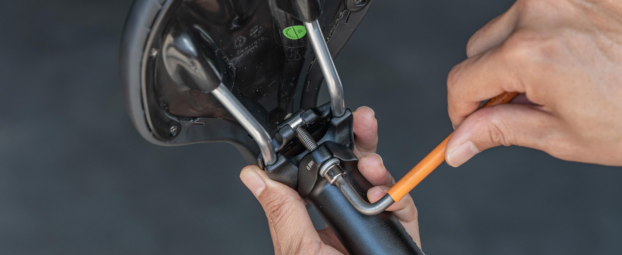 The image shows a close-up of a person using a hex key to adjust a saddle mounted on a seat post.slider_item_nEKeyV