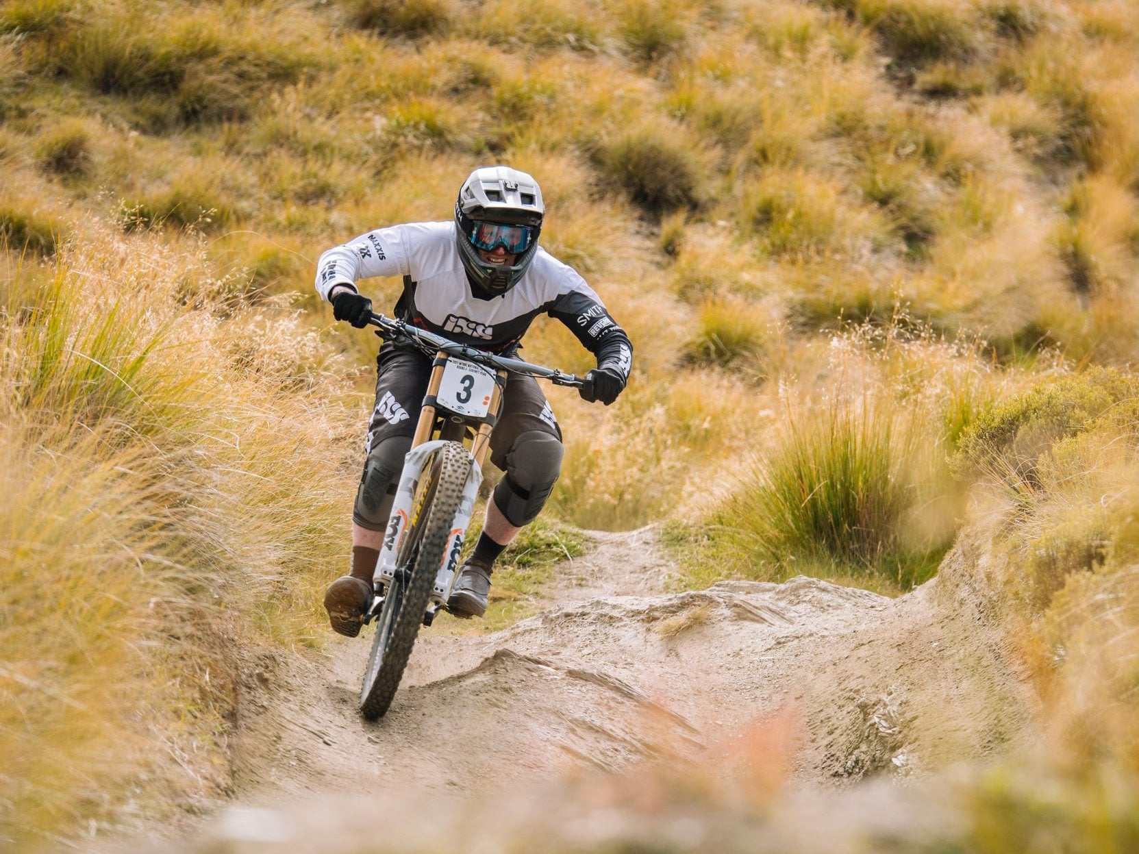 A mountain biker in full gear rides downhill on a rugged trail, surrounded by grassy terrain.